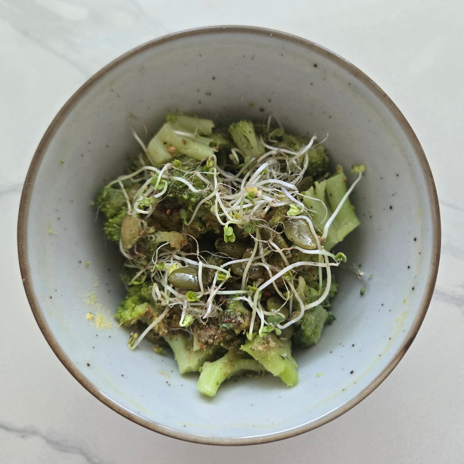 A bowl of cooked broccoli topped with raw broccoli sprouts, ground mustard powder, olive oil and raw pumpkin seeds