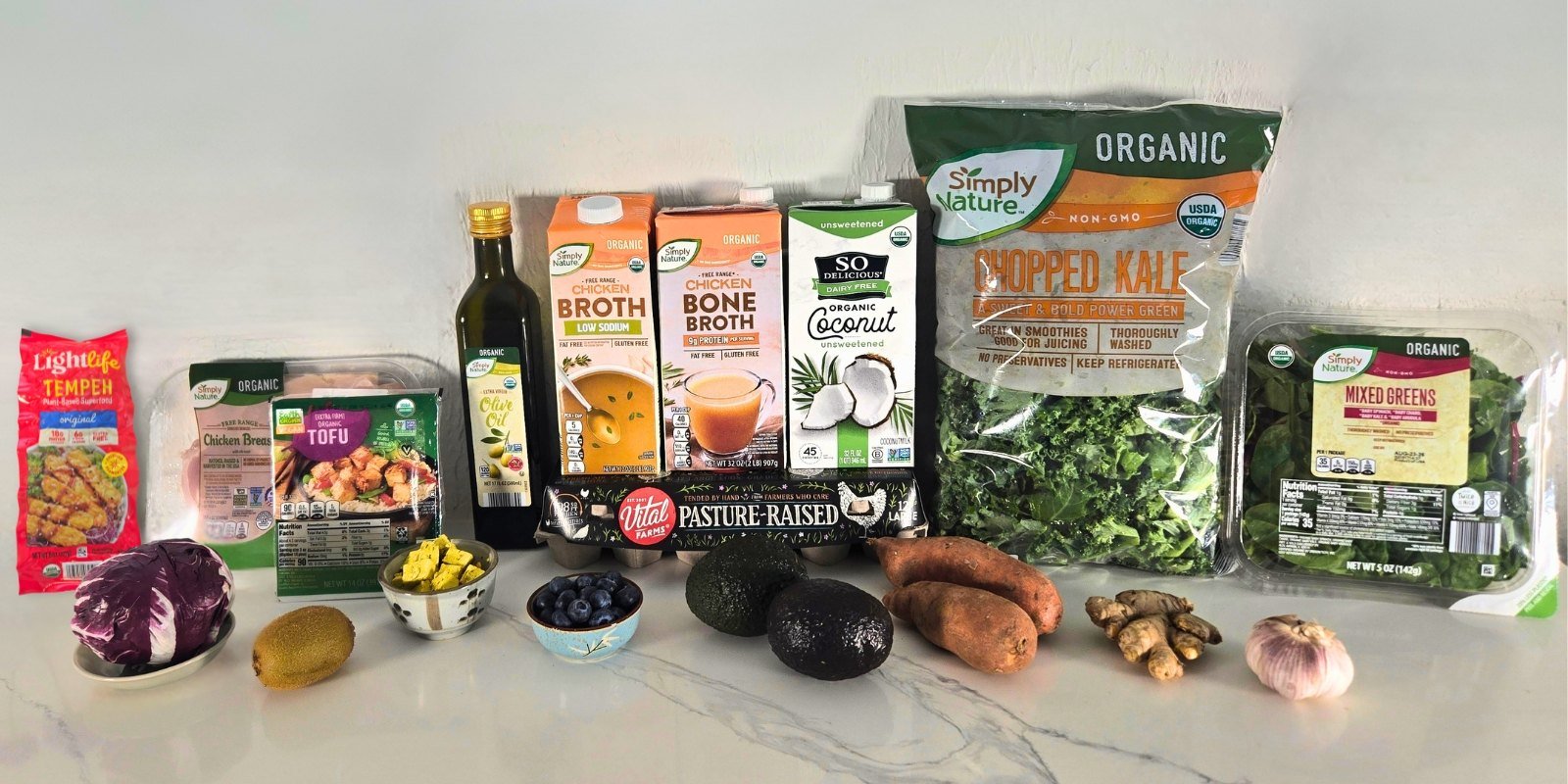 Organic groceries on a kitchen counter: Lightlife tempeh, Simply Nature free range chicken breast, organic extra firm tofu, Simply Nature organic olive oil, low sodium organic chicken broth, organic chicken bone broth, So Delicious unsweetened organic coconut milk, Simply Nature organic chopped kale and mixed greens, Vital Farms pasture-raised eggs, radicchio, kiwi, blueberries, avocados, sweet potatoes, fresh ginger and garlic.